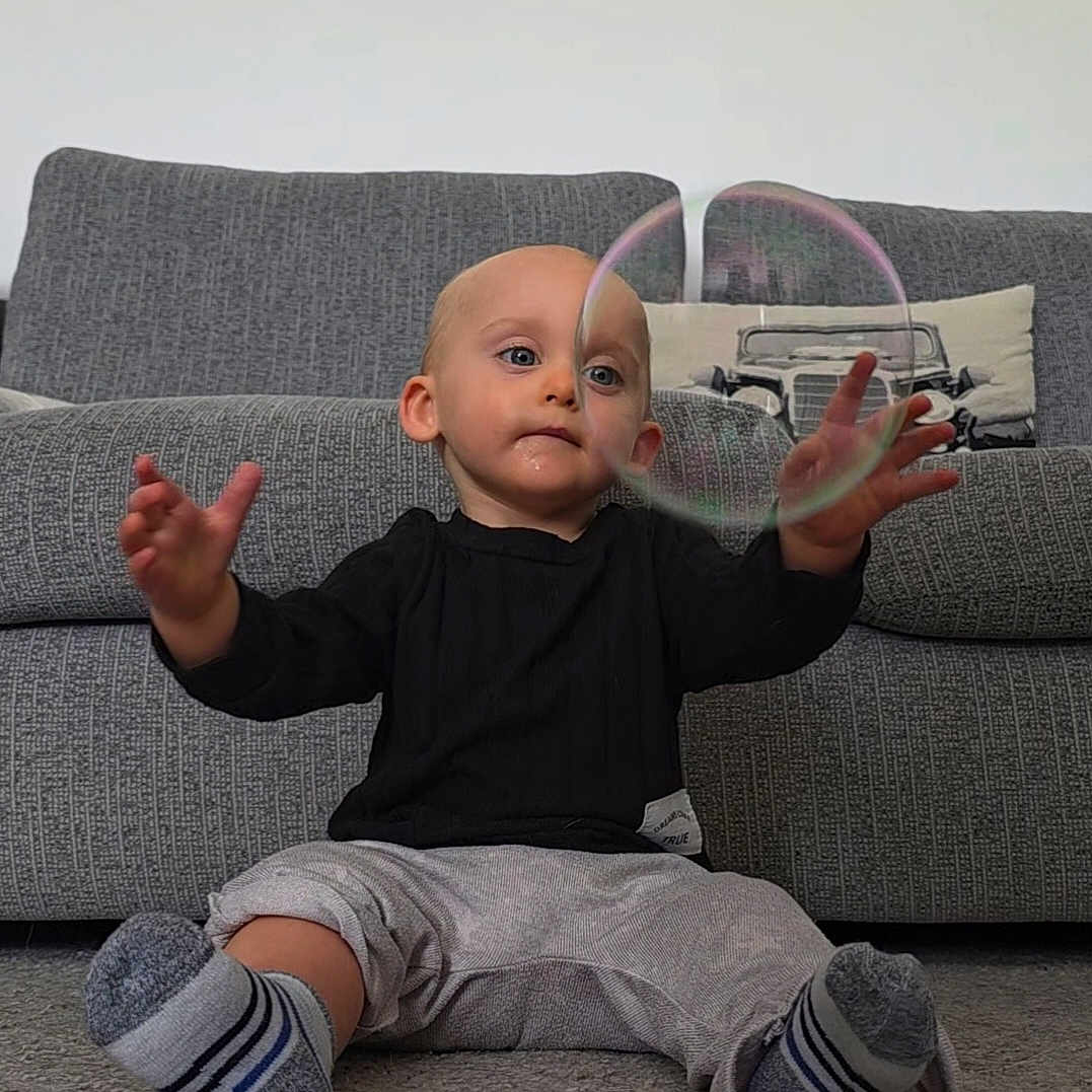 Vincent is registered to the contest to win money with this photo: baby, bubble, carpet, child, clothing, couch, curious, face, floor, hands, home, indoor, living_room, person, pillow, playing, reaching, soap_bubble, socks, toddler
