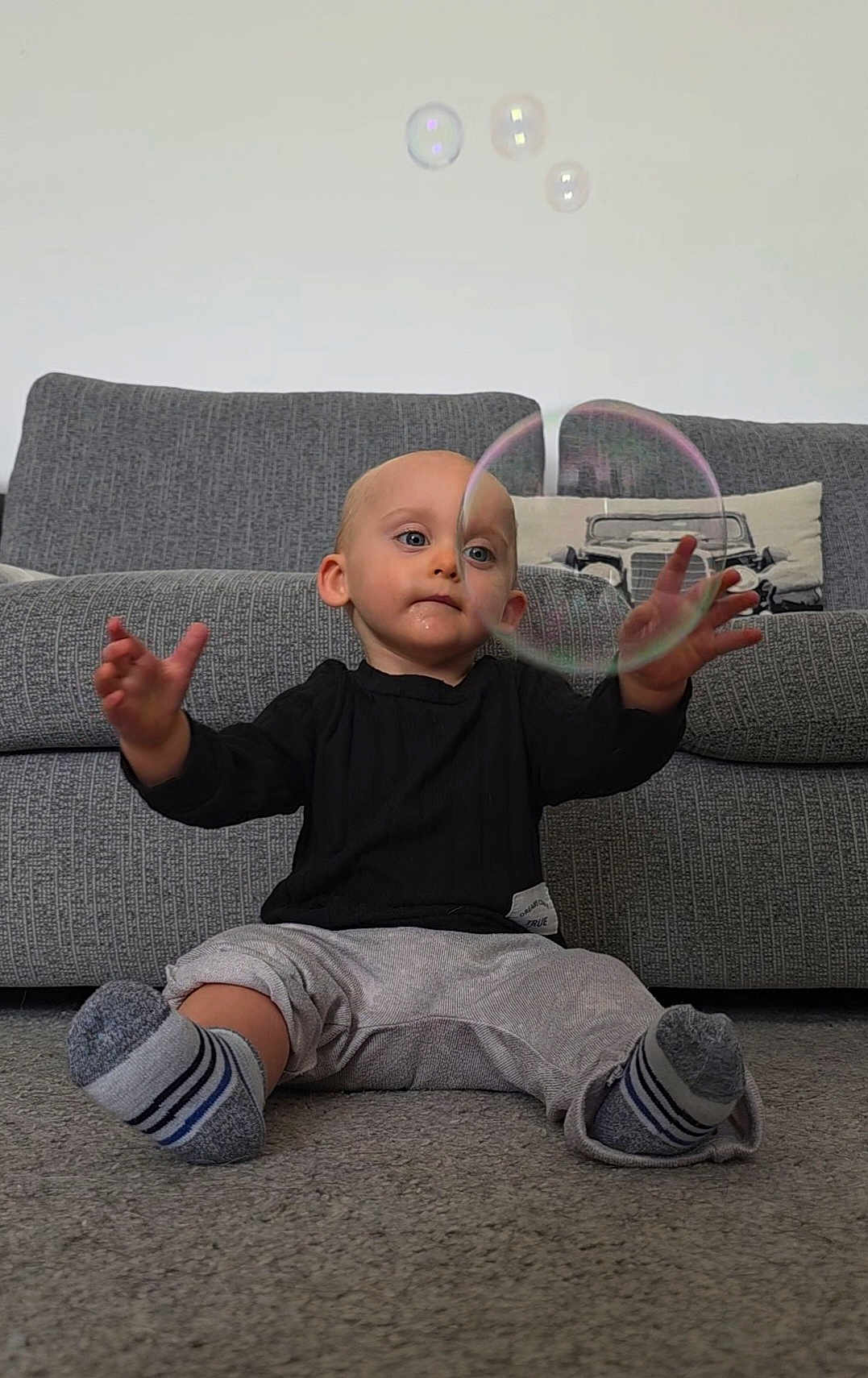 Vincent is registered to the contest to win money with this photo: toddler, child, bubble, soap_bubble, carpet, couch, pillow, indoor, socks, clothing, person, playing, curious, baby, hands, face, living_room, home, floor, reaching