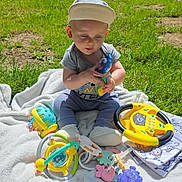 Vincent joined the competition — help win amazing prizes! baby, child, cap, toy, rattle, blanket, grass, outdoor, sunlight, play, steering_wheel, colorful, cute, sitting, infant, nature, park, happy, clothing, footwear