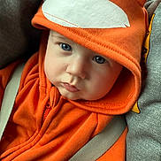 Carter is registered to the contest to win money with this photo: baby, child, hoodie, orange_clothing, fox_design, blue_eyes, car_seat, seatbelt, close_up, cute, infant, face, expression, soft_lighting, casual, warm_clothing, indoors, portrait, young_child, sitting