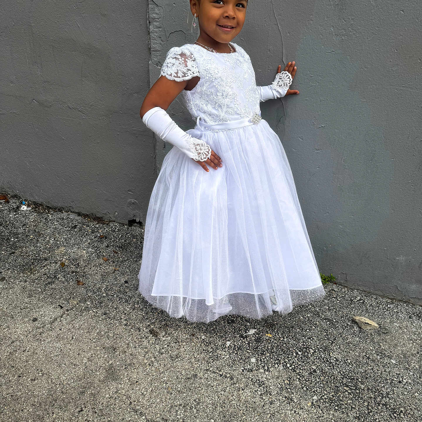 Athena is registered to the contest to win money with this photo: child, girl, white_dress, gloves, flower_crown, posing, smiling, outdoor, gray_wall, concrete_ground, fashion, portrait, young, elegant, special_occasion, lace, accessories, confident, cute, happy