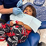 child, dental_chair, dentist, toy_truck, blue_gloves, dental_clinic, scrubs, medical_procedure, person, smiling, young_child, clean_environment, indoor, healthcare, plastic_cover, medical_equipment, white_shoes, red_clothing, black_pants, waiting