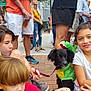 adult, bracelet, casual_clothing, child, costume, crowd, dinosaur_costume, dog, face, hair, leash, outdoor, pavement, people, shoes, shorts, sitting, smiling, street, tshirt