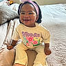 River is registered to the contest to win money with this photo: baby, child, smiling, headband, flower, tshirt, pink_shoes, pants, bed, blanket, indoor, cute, happy, person, portrait, sitting, young_child, clothing, footwear, home