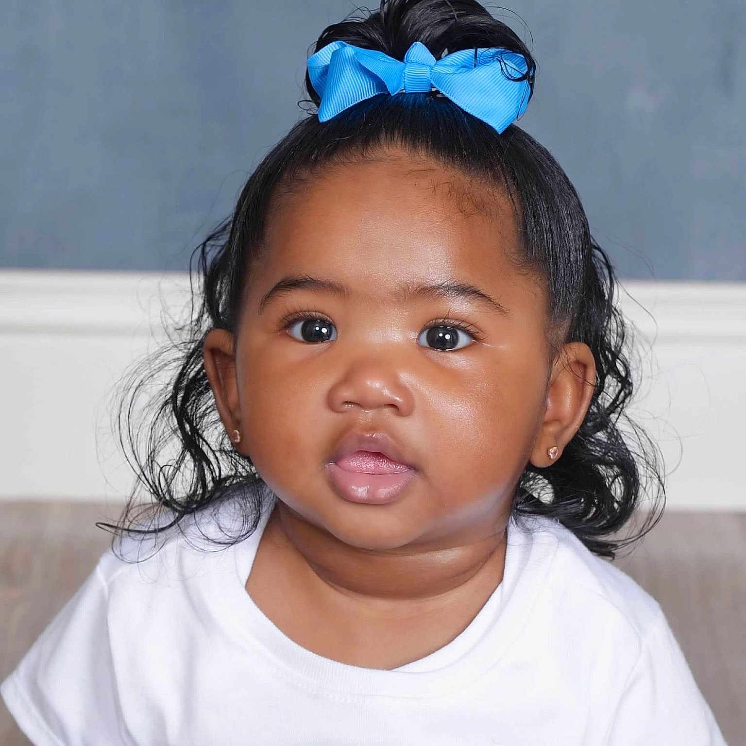 River is registered to the contest to win money with this photo: baby, child, portrait, face, blue_bow, white_shirt, curly_hair, cute, adorable, indoor, closeup, infant, young_child, skin, black_hair, eyes, expression, person, head, studio_lighting
