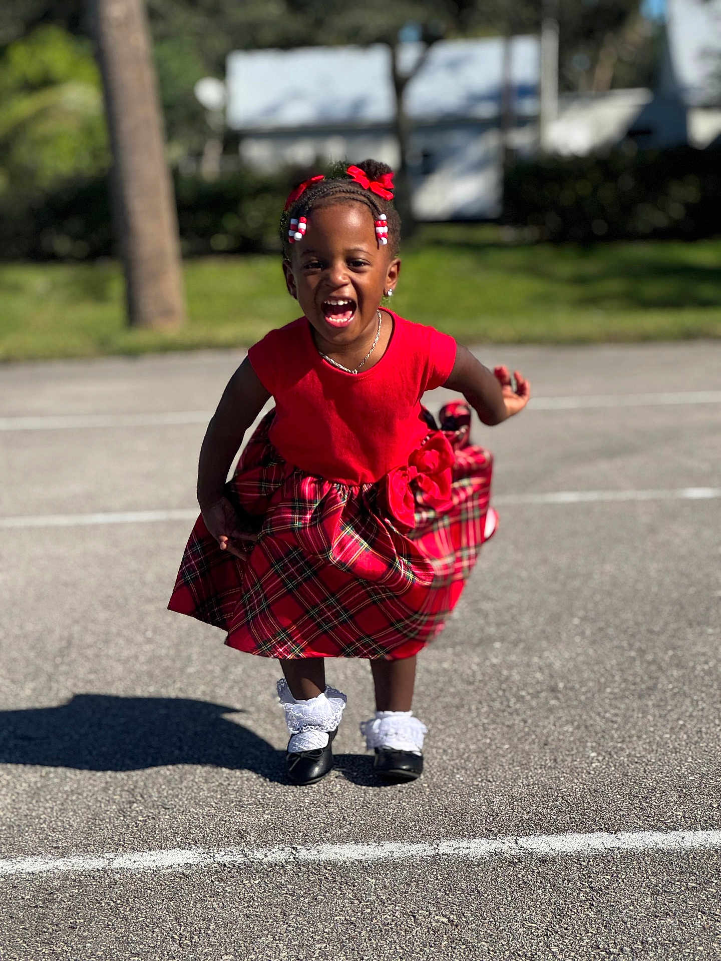 Nyomi joined the competition — help win amazing prizes! child, girl, red_dress, plaid_skirt, outdoor, laughing, smiling, sunny, pavement, shadow, black_shoes, white_socks, hair_bows, braids, necklace, earrings, greenery, tree, daylight, happy