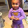 toddler, child, purple_outfit, indoor, floor, socks, cartoon_character, smile, standing, laundry_basket, messy_floor, curly_hair, person, cute, playful, home, clothing, expression, young, casual