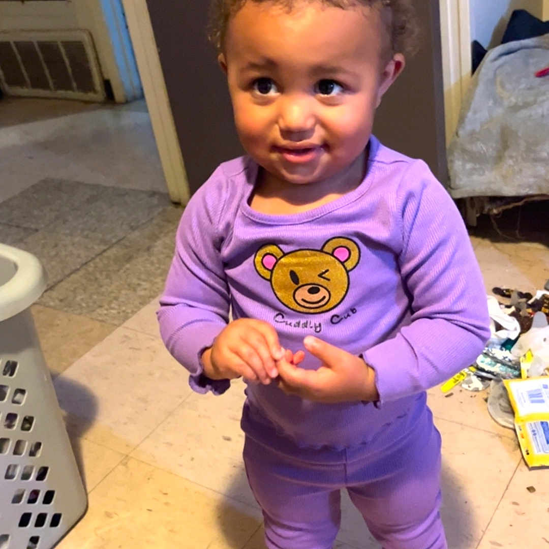Amariana is registered to the contest to win money with this photo: toddler, child, purple_outfit, indoor, floor, socks, cartoon_character, smile, standing, laundry_basket, messy_floor, curly_hair, person, cute, playful, home, clothing, expression, young, casual