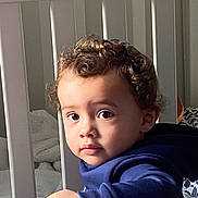 Kyron joined the competition — help win amazing prizes! toddler, child, curly_hair, face, eyes, crib, blanket, hoodie, indoor, portrait, baby, hand, expression, furniture, person, young_child, cozy, soft_light, curious, skin