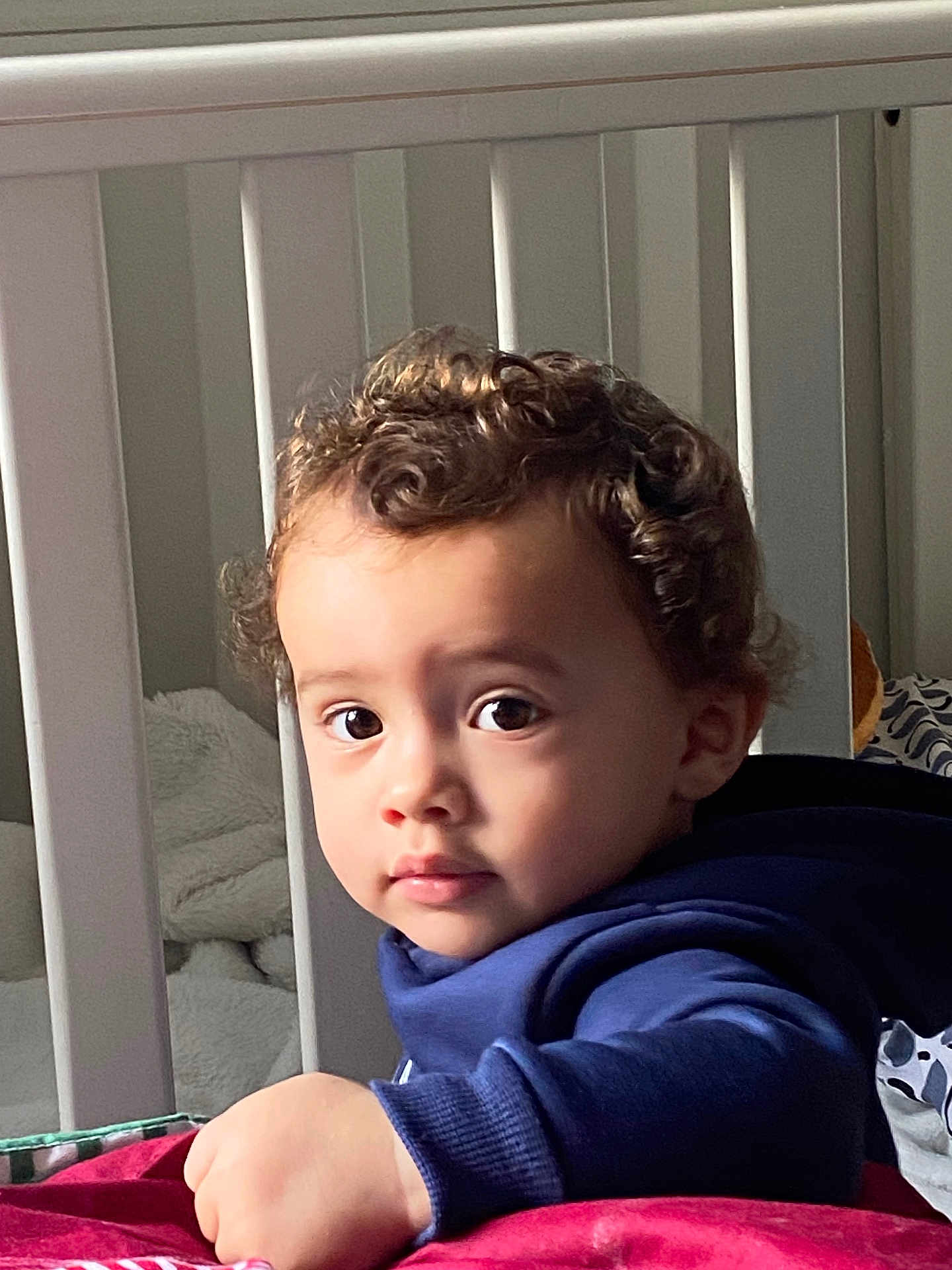 Kyron joined the competition — help win amazing prizes! toddler, child, curly_hair, face, eyes, crib, blanket, hoodie, indoor, portrait, baby, hand, expression, furniture, person, young_child, cozy, soft_light, curious, skin