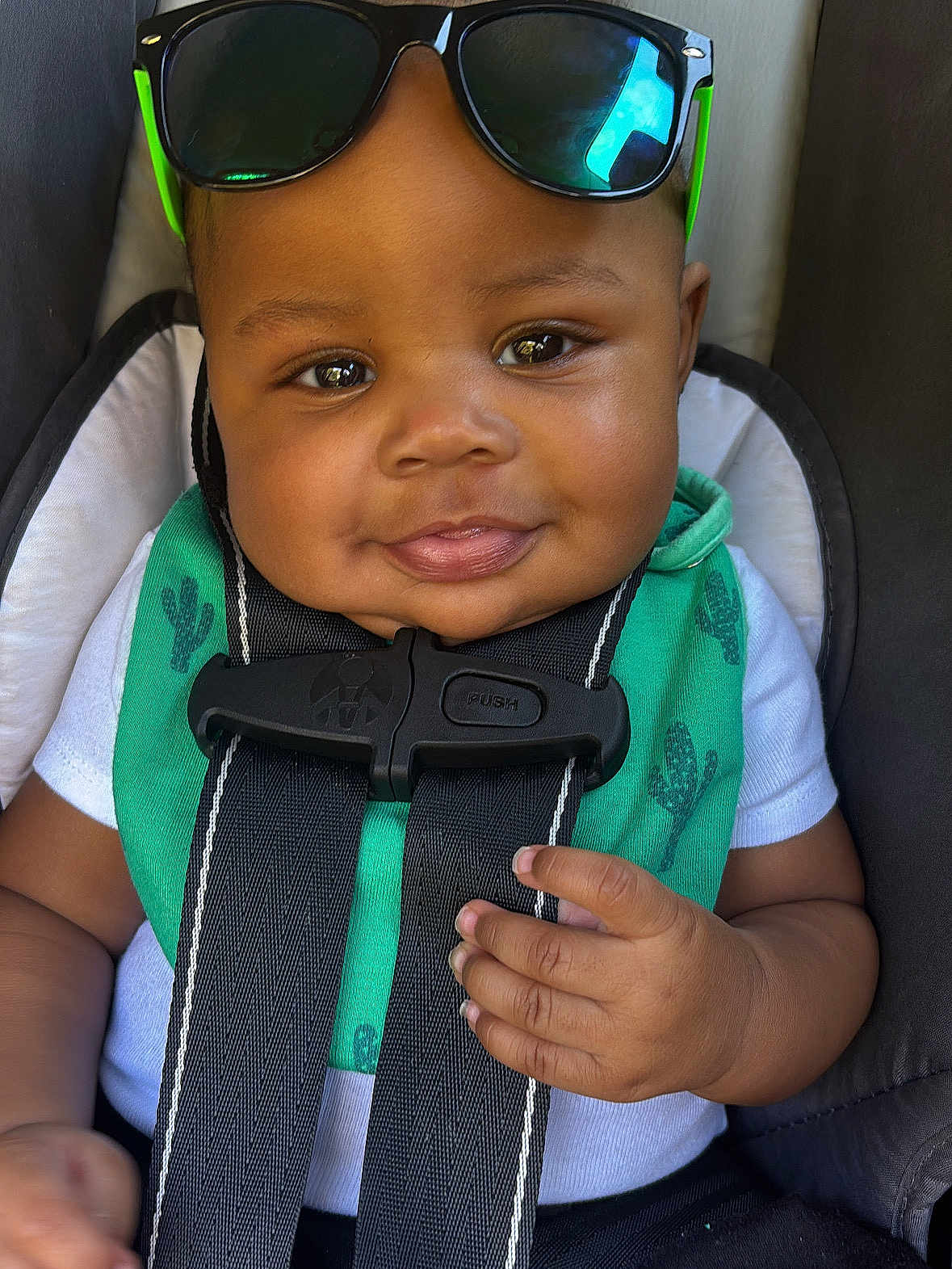 Nova joined the competition — help win amazing prizes! baby, child, car_seat, sunglasses, smile, face, hand, clothing, bib, seatbelt, infant, cute, portrait, person, green, black, white, cactus, safety, closeup