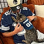 person, cat, tabby_cat, sports_jersey, baseball_cap, couch, pillow, indoor, tattoo, casual_clothing, pet, holding, seated, brown_furniture, gray_pants, affection, animal_clothing, relaxation, human_animal_interaction, domestic