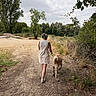 person, dog, leash, walking, dirt_path, trees, grass, shrubs, outdoor, nature, cloudy_sky, casual_clothing, footwear, summer, back_view, animal, walking_path, greenery, daytime, scenic
