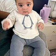 Samuel is registered to the contest to win money with this photo: baby, child, sweater, penguin, knitted_pants, cushion, indoor, furniture, water_bottle, lamp, person, seated, curious, wide_eyes, hand, face, clothing, home, bedside_table, purple