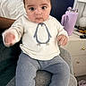 baby, child, sweater, penguin, knitted_pants, cushion, indoor, furniture, water_bottle, lamp, person, seated, curious, wide_eyes, hand, face, clothing, home, bedside_table, purple