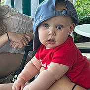Philip is registered to the contest to win money with this photo: baby, child, cap, red_shirt, blue_cap, curious_expression, chubby_arms, adult_hand, outdoor, greenery, towel, seated, person, casual, skin, table, chair, daylight, portrait, hands