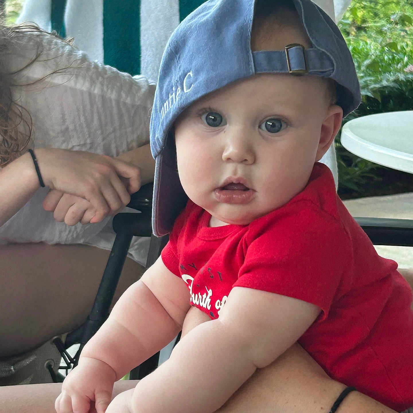 Philip is registered to the contest to win money with this photo: adult_hand, baby, blue_cap, cap, casual, chair, child, chubby_arms, curious_expression, daylight, greenery, hands, outdoor, person, portrait, red_shirt, seated, skin, table, towel