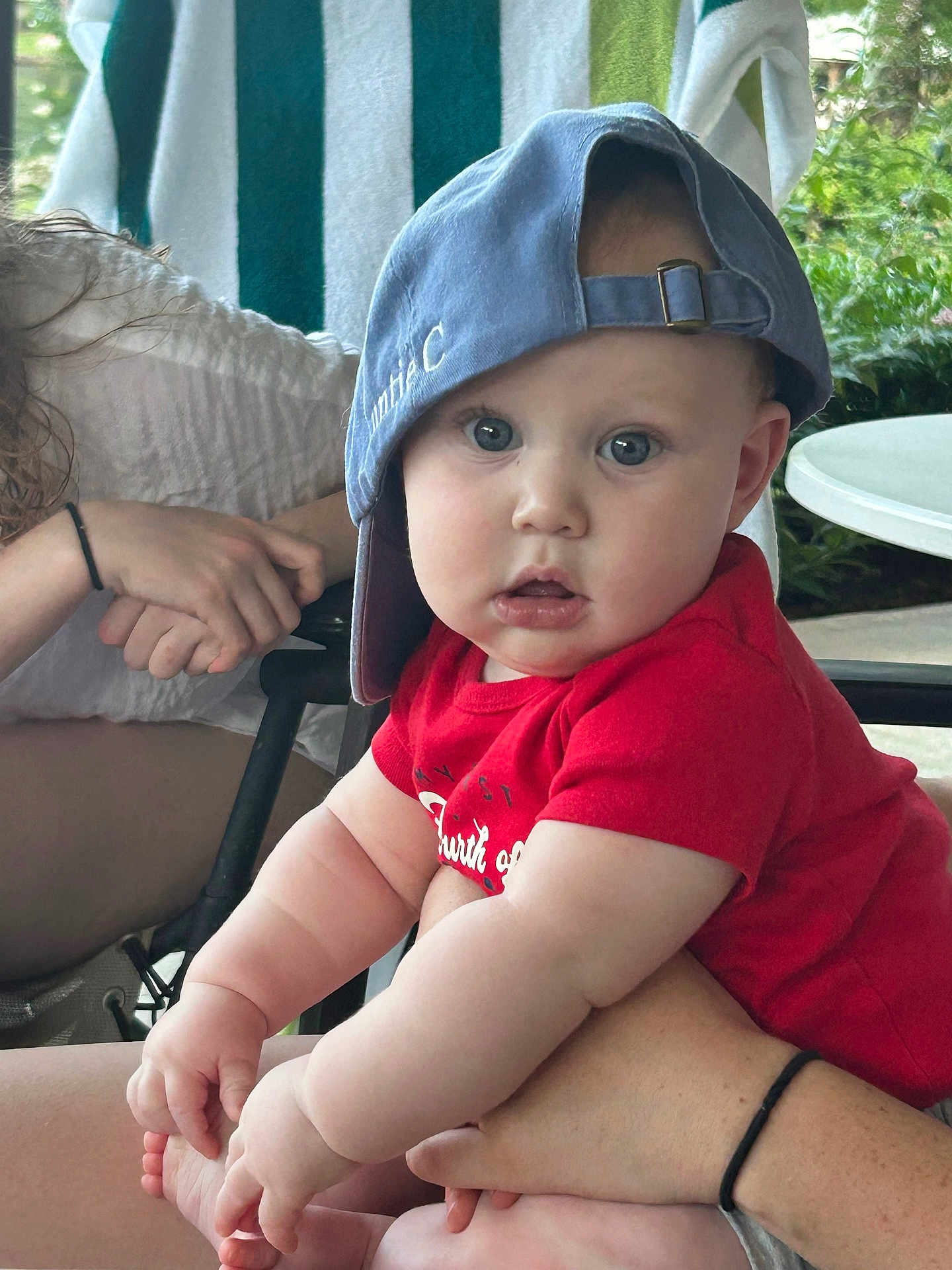 Philip is registered to the contest to win money with this photo: baby, child, cap, red_shirt, blue_cap, curious_expression, chubby_arms, adult_hand, outdoor, greenery, towel, seated, person, casual, skin, table, chair, daylight, portrait, hands