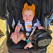 Letty is registered to the contest to win money with this photo: baby, child, stroller, headband, fan, foot, sunlight, outdoor, smiling, cute, clothing, playful, seatbelt, person, infant, summer, accessory, happy, black, orange