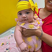 Abryana joined the competition — help win amazing prizes! baby, child, infant, yellow_headband, pink_outfit, sun_pattern, adult_hand, smile, yellow_wall, blanket, cartoon_characters, indoor, holding, cute, portrait, person, clothing, headwear, happy, toddler