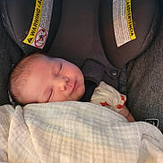 Tobias is registered to the contest to win money with this photo: baby, sleeping, blanket, car_seat, child, infant, face, head, clothing, warning_label, seatbelt, soft_texture, rest, peaceful, closeup, portrait, indoors, comfort, newborn, cute