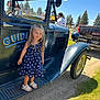 child, girl, vintage_truck, blue_dress, sandals, outdoor, sunny, grass, trees, sky, vehicle, smile, standing, daylight, classic_car, nature, people, park, reflection, side_view