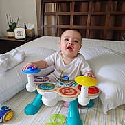 Sebastian joined the competition — help win amazing prizes! baby, child, bed, toy, musical_toy, onesie, indoor, furniture, headboard, curtains, plant, tissue_box, photo_frame, expression, playing, colorful, person, smiling, sitting, room