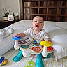 baby, child, bed, toy, musical_toy, onesie, indoor, furniture, headboard, curtains, plant, tissue_box, photo_frame, expression, playing, colorful, person, smiling, sitting, room