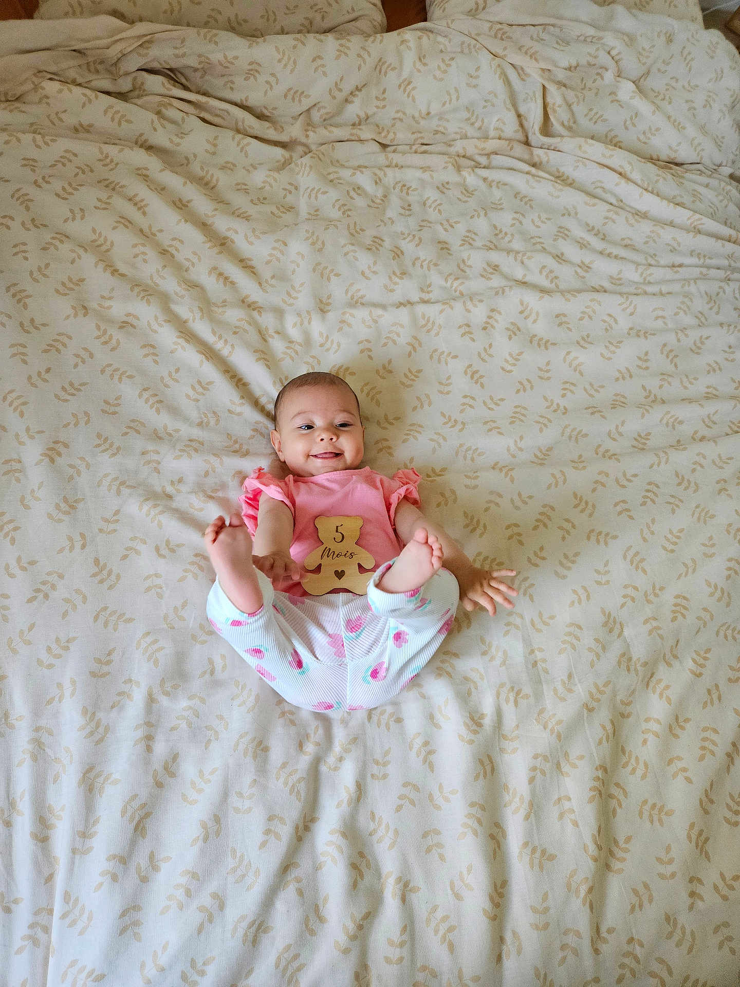 Irina participe au concours pour gagner de l'argent avec cette photo : baby, infant, smiling, lying_down, pink_shirt, white_pants, patterned_bedspread, wooden_sign, feet_up, cute, child, person, indoors, bed, fabric, happy, young, portrait, human, sleepwear