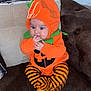 baby, child, costume, couch, cute, face, funny, furniture, halloween, hands, holiday, indoor, infant, orange_clothing, person, pillows, plush, pumpkin_costume, sitting, striped_pants