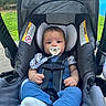 baby, pacifier, car_seat, blue_clothing, knitwear, infant, child, outdoor, greenery, curious, sitting, straps, safety_harness, face, head, white_socks, cute, portrait, young_child, daylight