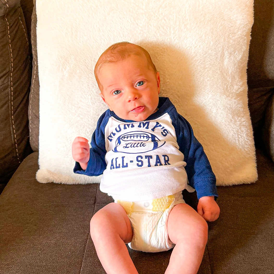 Kamden is registered to the contest to win money with this photo: baby, blue_eyes, brown_couch, casual, clothing, couch, cute, diaper, fabric, furniture, indoor, infant, person, pillow, portrait, raised_fist, sitting, smile, white_pillow, young_child