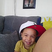 Terrance Jr is registered to the contest to win money with this photo: child, smiling, portrait, indoor, couch, sofa, cap, hat, yellow_shirt, family, home, living_room, framed_photo, wall, cushion, happy, teeth, sitting, close_up, photography