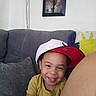 child, smiling, portrait, indoor, couch, sofa, cap, hat, yellow_shirt, family, home, living_room, framed_photo, wall, cushion, happy, teeth, sitting, close_up, photography
