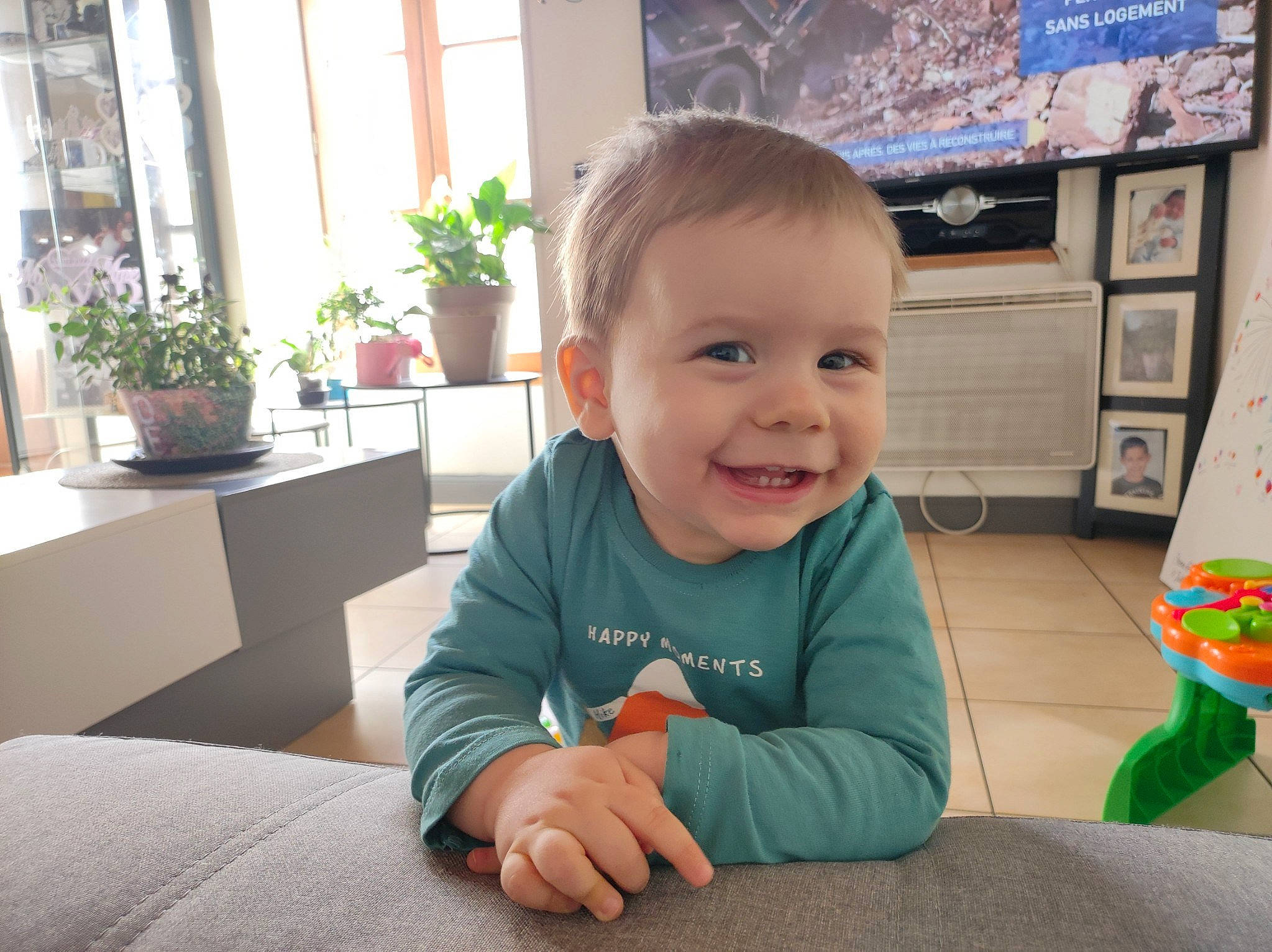 Elyo participe au concours pour gagner de l'argent avec cette photo : baby, baby_toddler_clothing, child, flooring, flowerpot, fun, happy, houseplant, joy, person, plant, room, sitting, skin, smile, standing, table, television, toddler, tree