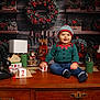 baby, child, christmas, holiday, festive_clothing, elf_outfit, table, wooden_table, wreath, candles, teddy_bear, nutcracker, vintage_telephone, decorations, red_berries, greenery, smiling, indoors, hat, jeans