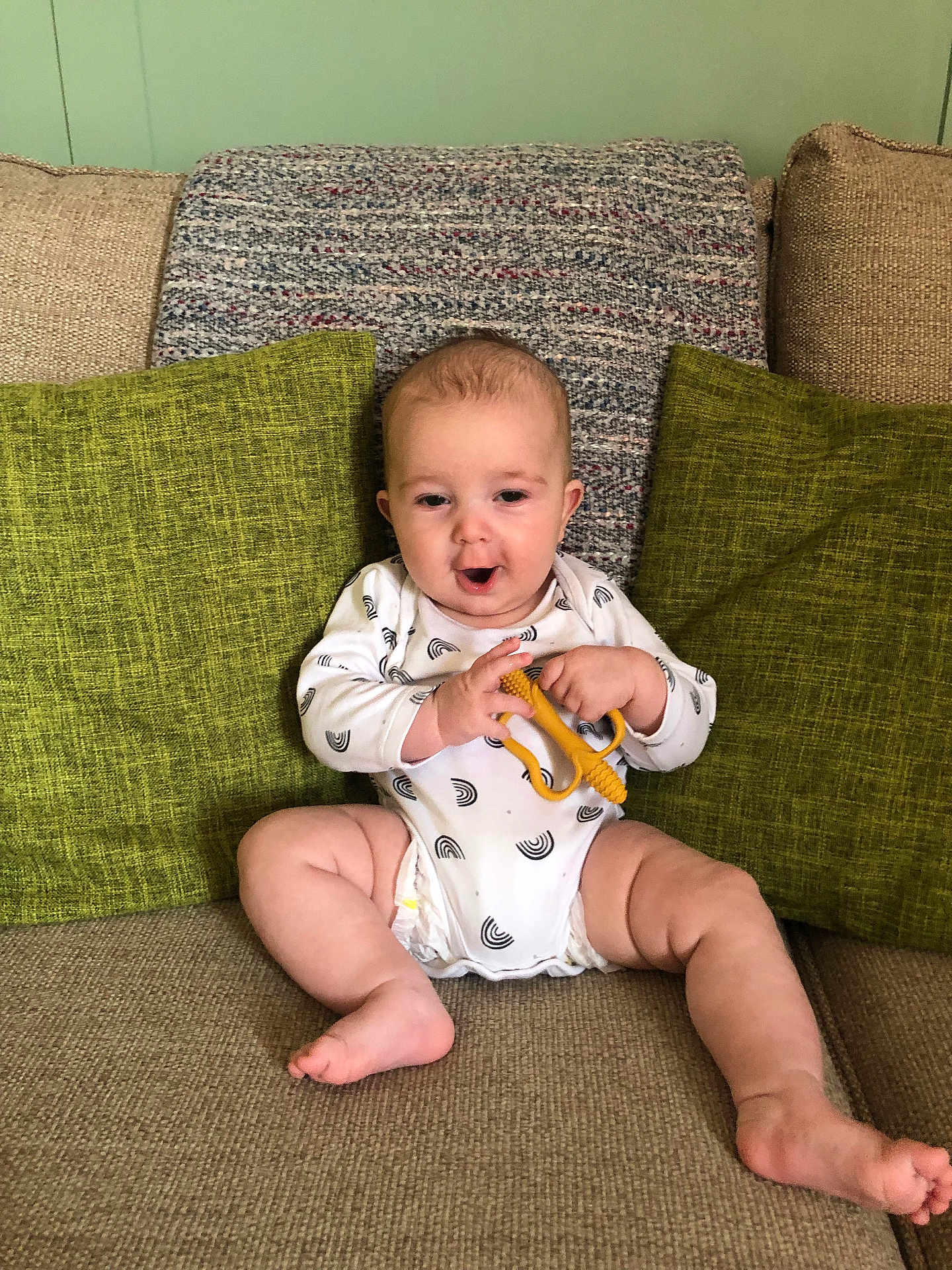 Finley is registered to the contest to win money with this photo: baby, infant, child, couch, pillow, green_pillow, onesie, teething_toy, beige_couch, indoor, sitting, cute, chubby, expression, holding, furniture, home, smiling, face, person