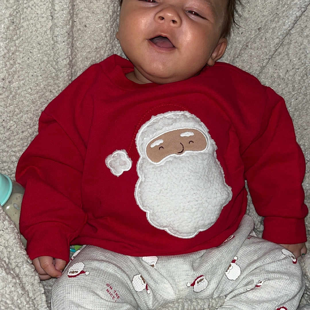 Demonta is registered to the contest to win money with this photo: baby, blanket, child, christmas, cozy, cute, face, happy, holiday, indoors, infant, pajamas, patterned_pants, person, red_sweater, santa_claus, sitting, smiling, soft_texture, warm