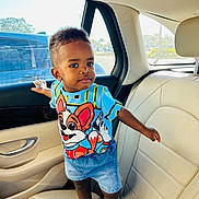 Chris Roy is registered to the contest to win money with this photo: car_door, car_interior, cartoon_shirt, child, clothing, curious, daylight, face, footwear, hand, head, leather_seat, person, sandals, seatbelt, shorts, standing, toddler, window, young_child