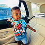 car_door, car_interior, cartoon_shirt, child, clothing, curious, daylight, face, footwear, hand, head, leather_seat, person, sandals, seatbelt, shorts, standing, toddler, window, young_child