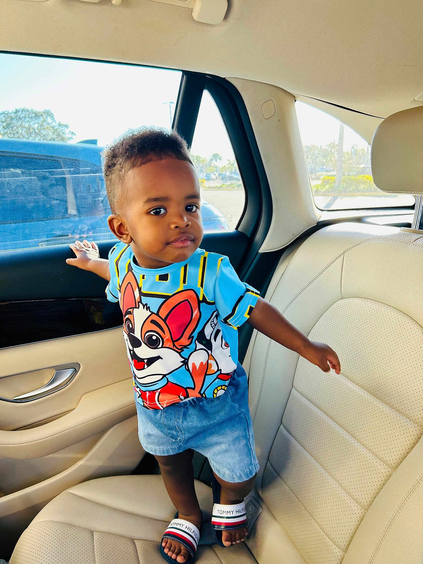 Chris Roy is registered to the contest to win money with this photo: car_door, car_interior, cartoon_shirt, child, clothing, curious, daylight, face, footwear, hand, head, leather_seat, person, sandals, seatbelt, shorts, standing, toddler, window, young_child