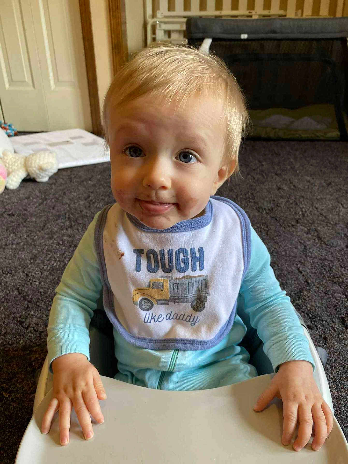 Ryan is registered to the contest to win money with this photo: toddler, child, baby, bib, high_chair, smiling, face, indoor, carpet, toy, clothing, hand, expression, blonde_hair, sitting, cute, messy_face, person, young_child, playful