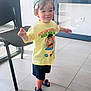 toddler, child, indoor, standing, cap, yellow_tshirt, graphic_tshirt, shorts, sandals, smiling, floor, tile_floor, chair, glass_door, natural_light, person, casual_clothing, young_child, playful, happy
