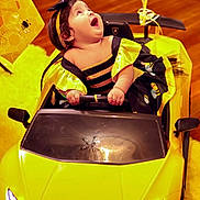 Kailani is registered to the contest to win money with this photo: baby, child, costume, bumblebee, toy_car, lamborghini, yellow, indoor, floor, headband, happy, smiling, hands, seat, vehicle, flooring, decorations, face, person, cute