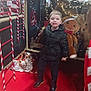 child, boy, black_jacket, boots, red_carpet, gingerbread_man, holiday_decorations, candy_cane, fairy_lights, festive, indoor, smiling, toy, christmas, decoration, holiday, display, seasonal, celebration, winter_clothing