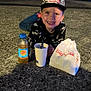 child, boy, smiling, cap, marvel, puma, jeans, bottle, drink, cup, straw, paper_bag, outdoor, night, pavement, happy, casual, food, person, evening