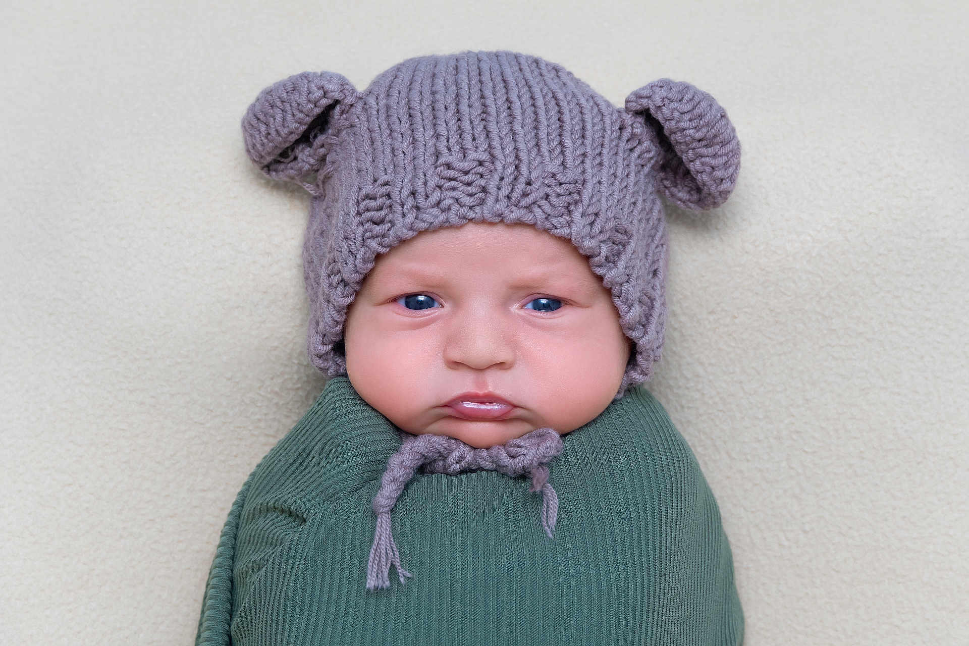 Richard is registered to the contest to win money with this photo: baby, infant, knitted_hat, bear_ears, blue_eyes, swaddle, green_clothing, chubby_cheeks, portrait, close_up, cute, serious_expression, soft_background, texture, headwear, child, newborn, indoors, warm_clothing, young_child