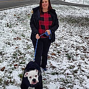 Daisy is registered to the contest to win money with this photo: woman, dog, leash, snow, grass, winter_clothing, jacket, road, sidewalk, house, tree, outdoor, pet, walking, cold_weather, smiling, black_pants, white_dog, red_checkered_shirt, park