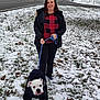 woman, dog, leash, snow, grass, winter_clothing, jacket, road, sidewalk, house, tree, outdoor, pet, walking, cold_weather, smiling, black_pants, white_dog, red_checkered_shirt, park
