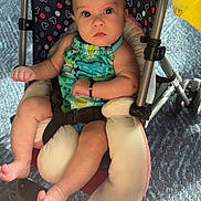 Charlotte is registered to the contest to win money with this photo: baby, child, infant, stroller, sitting, portrait, face, wide_eyed, looking_at_camera, colorful_clothing, romper, barefoot, feet, carpet, indoor, patterned_fabric, padded_seat, harness, wheelchair_like_stroller, photography